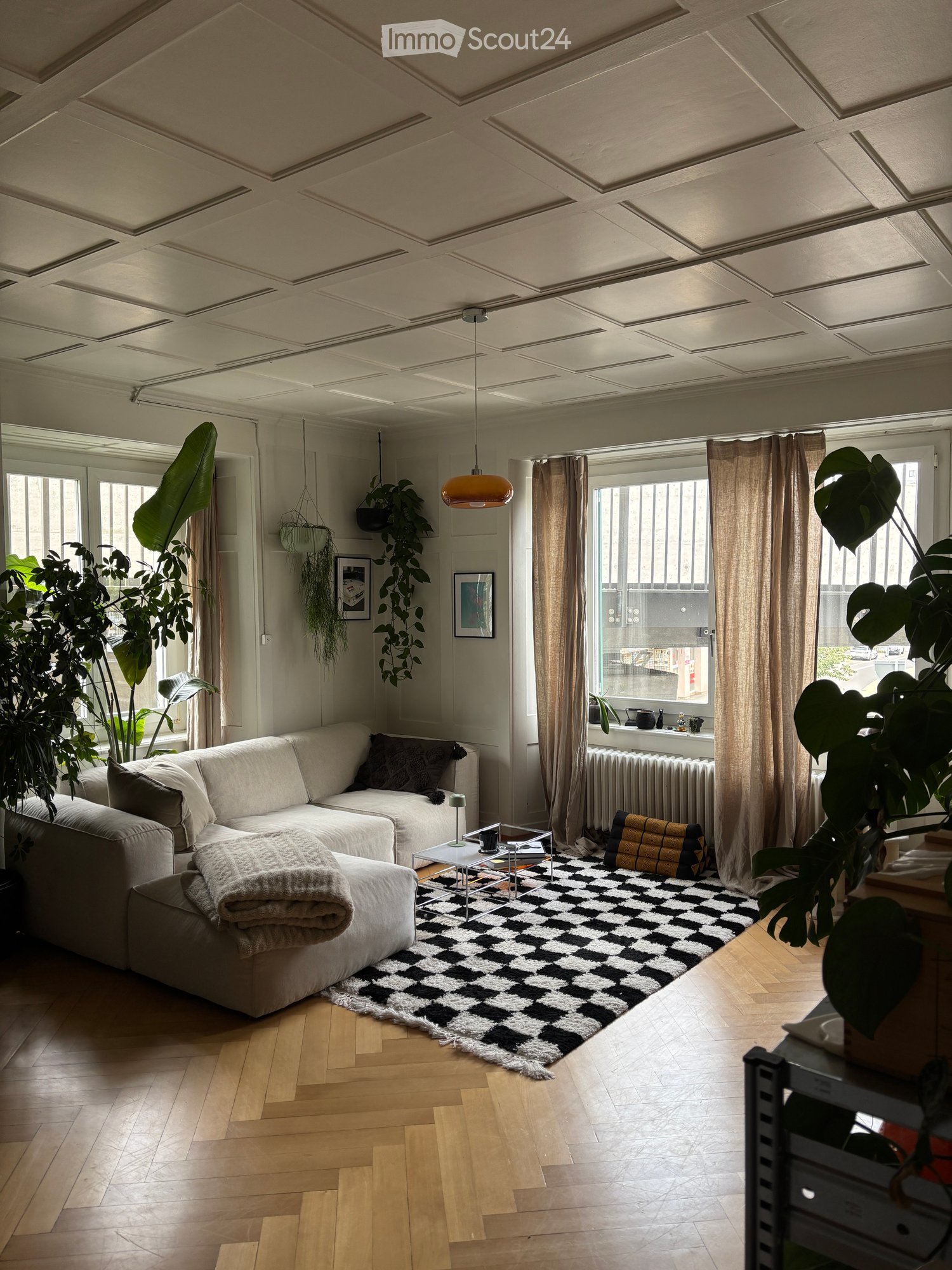 Modern living room with light-colored sofa, coffee table, floor lamp, and large potted plants. Features a black and white checkered rug, wooden flooring, and large windows with curtains.