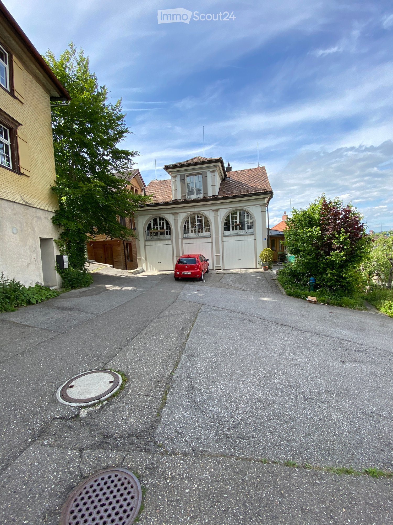 A two-story residential building with a garage, large windows, and a driveway with a red car parked in front.