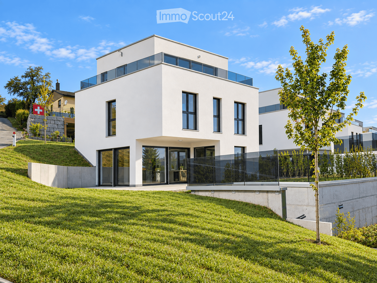 Modern two-story white house with large windows and a raised ground floor, surrounded by a well-maintained lawn and trees.