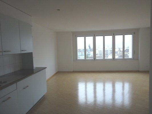 Empty room with wooden flooring, white walls, and a fitted kitchen area with cabinets and a countertop. Large windows provide natural light and a view of the city.