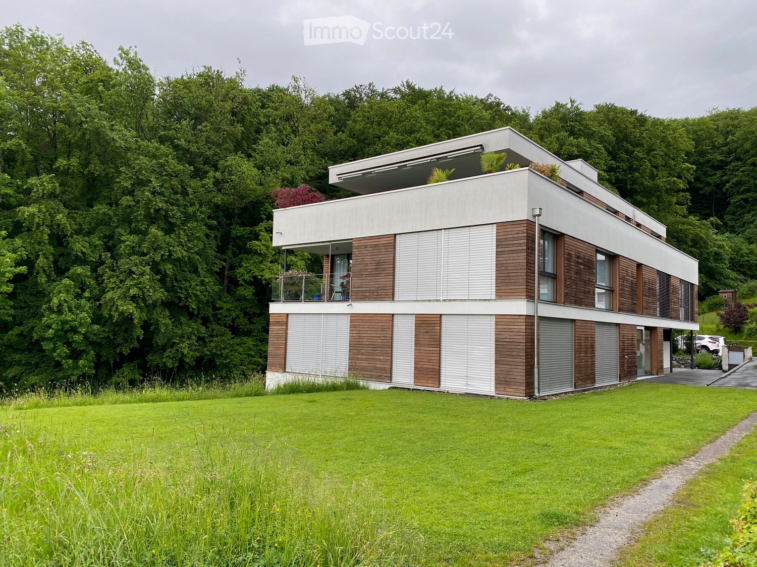 Modern two-story house with white and wood exterior, large windows, balcony, and surrounding greenery.