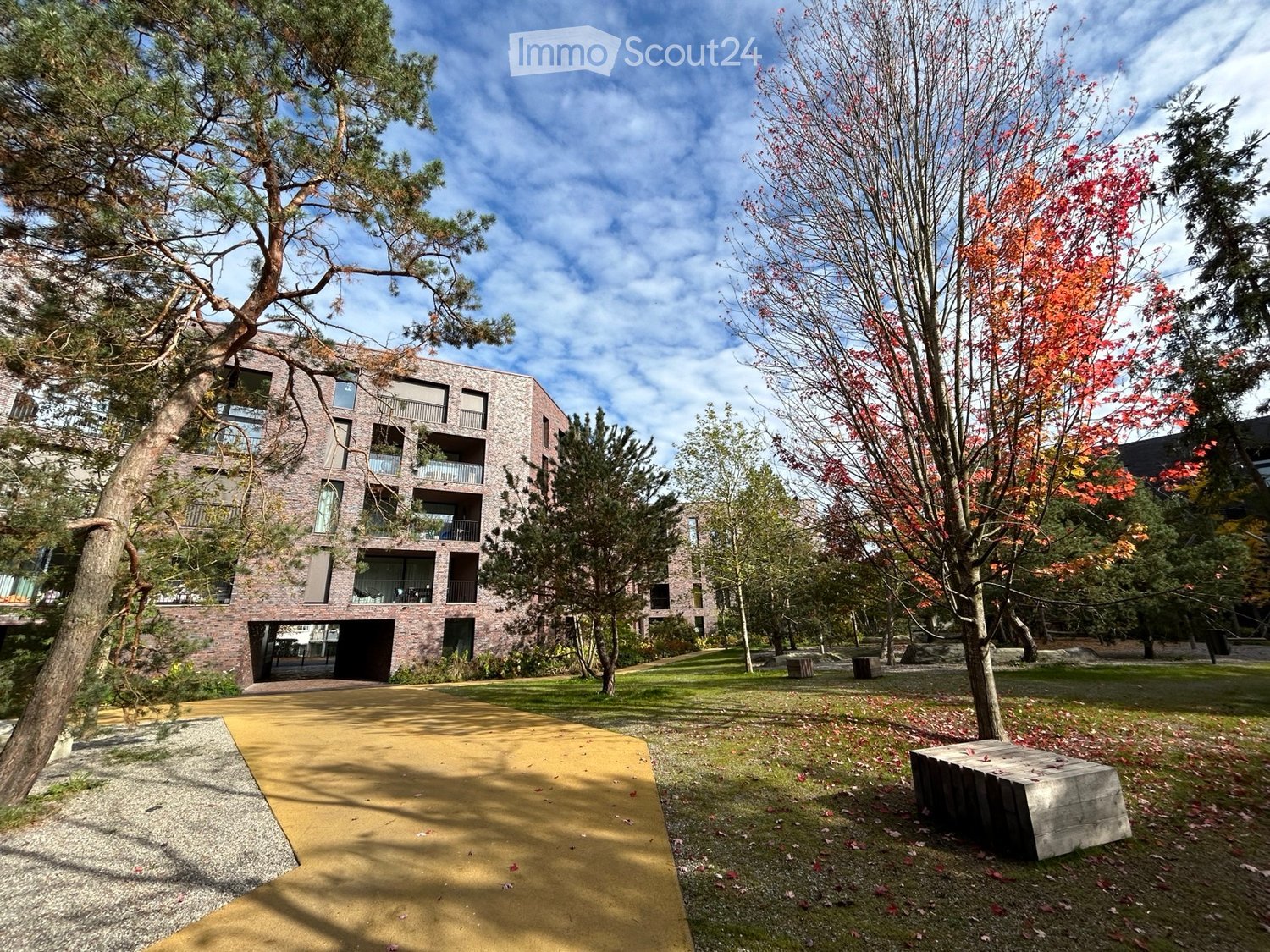 Modern brick building with multiple floors, balconies, and large windows, surrounded by trees and a paved walkway.
