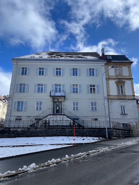 A large, multi-story building with white walls, multiple windows, and a staircase leading to the entrance. Snow is visible on the ground and roof.