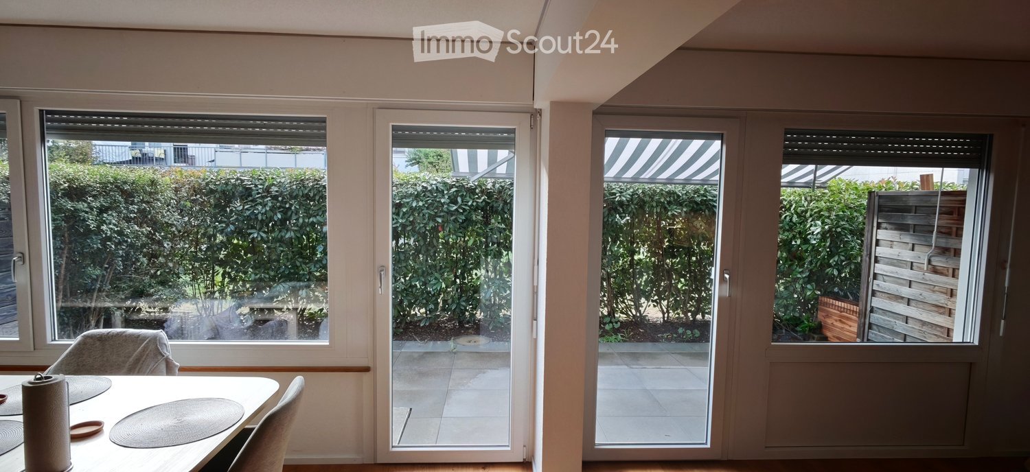 Bright dining area with large windows and sliding glass doors leading to a balcony or terrace. The room features a dining table with chairs and a view of a well-maintained garden.