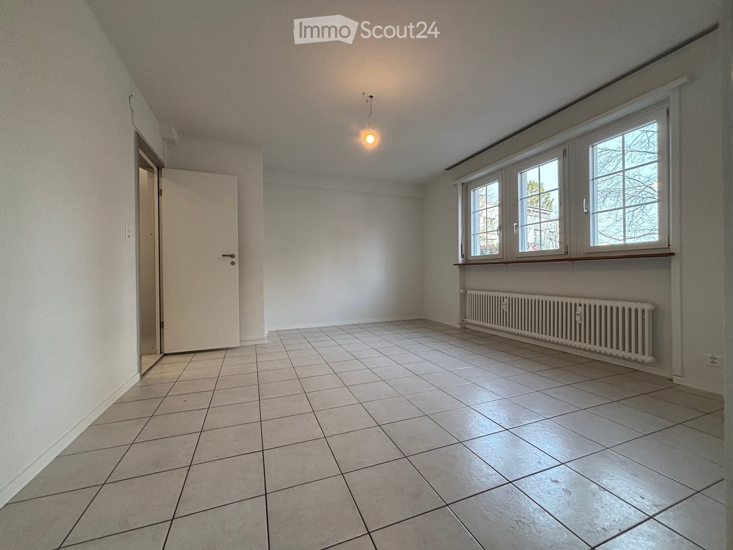Empty room with tiled floor, white walls, a window with blinds, a radiator, and a door leading to another space.