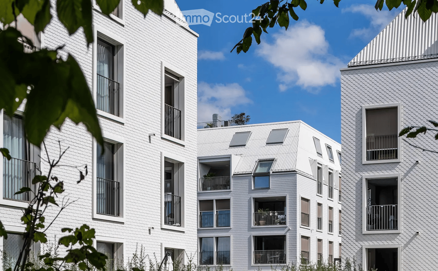 Modern white apartment building with multiple balconies and large windows, surrounded by greenery.