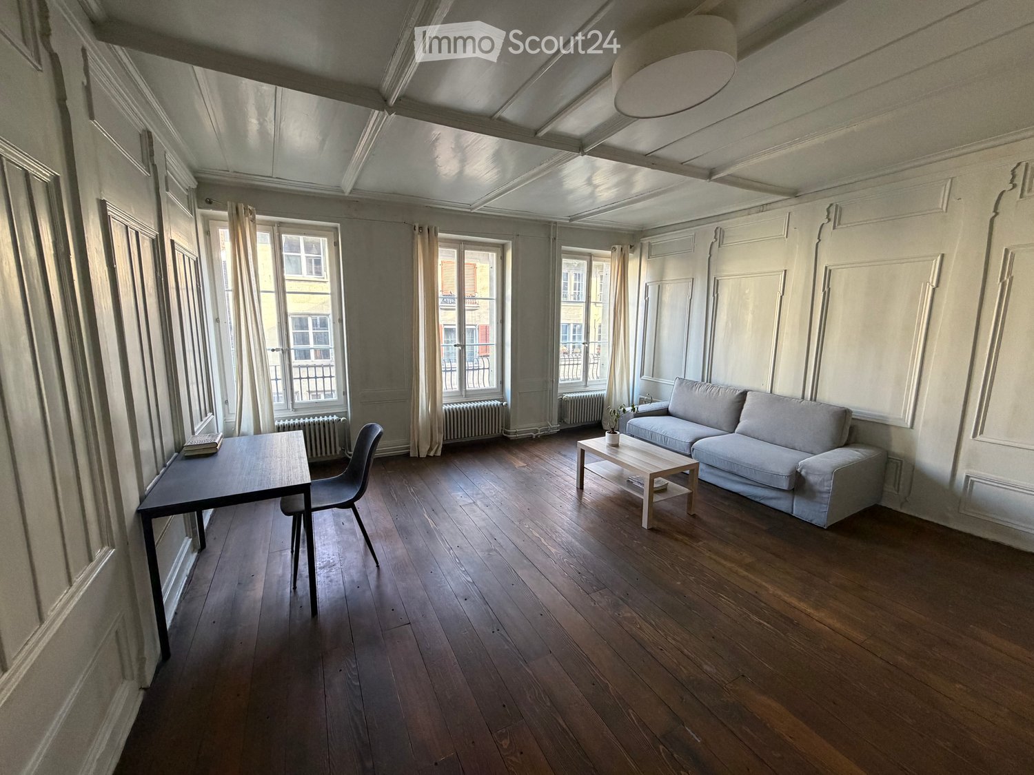 Living room with wooden flooring, large windows, a sofa, coffee table, and a small table with a chair.
