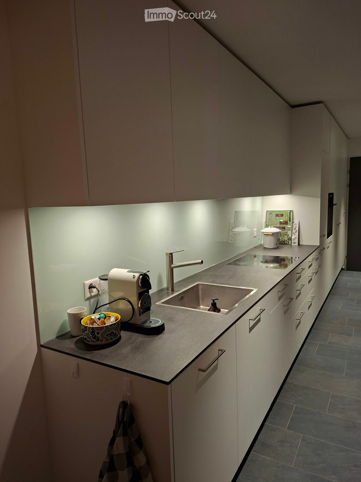 Modern kitchen with white cabinets, a stainless steel sink, and a coffee machine on the counter. The space features a sleek design with built-in lighting and a reflective backsplash.