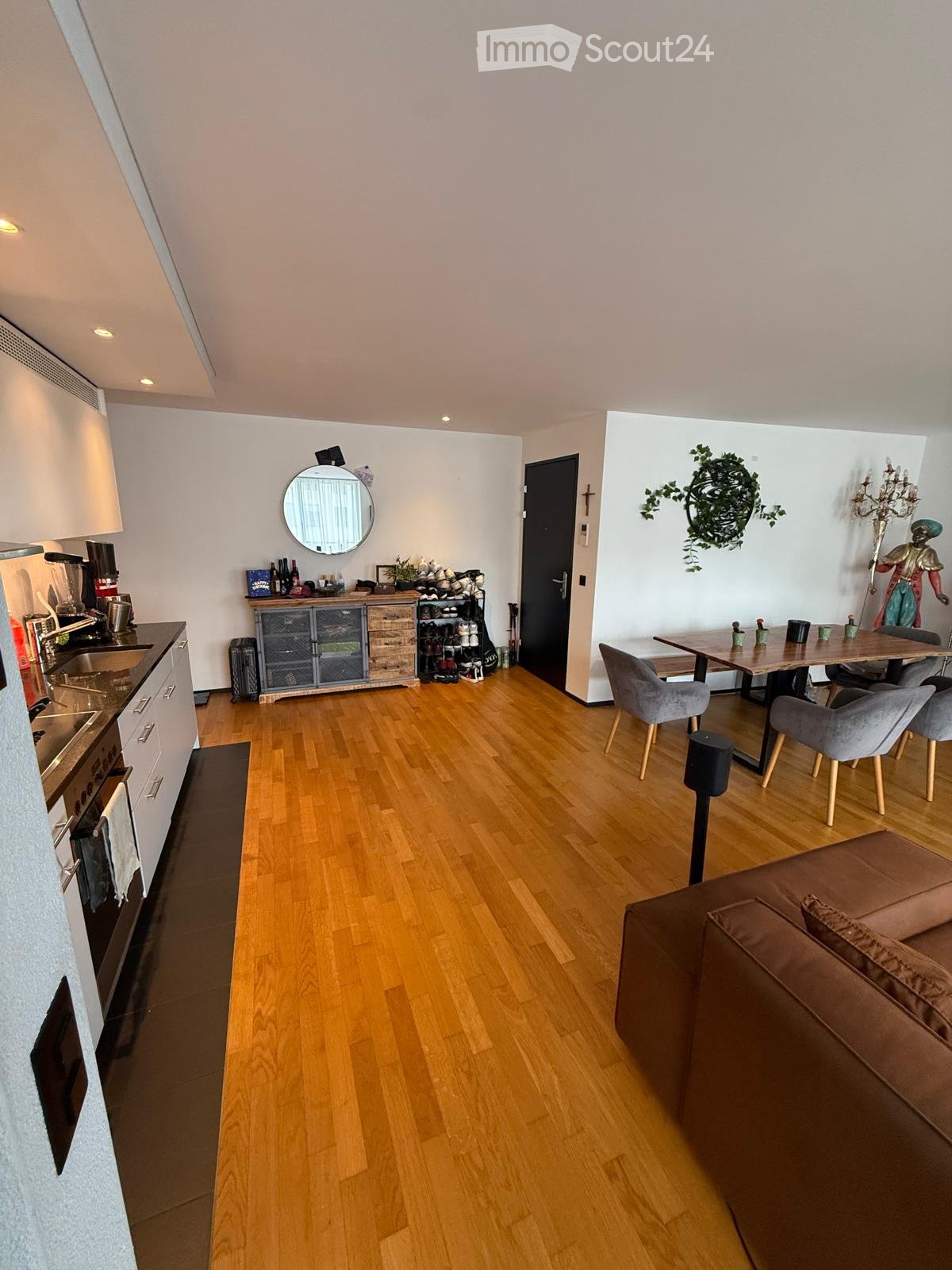 Open-plan living and dining area with wooden flooring, a brown sofa, a dining table with chairs, a kitchen area with cabinets, sink, stove, and a shoe rack near the entrance.