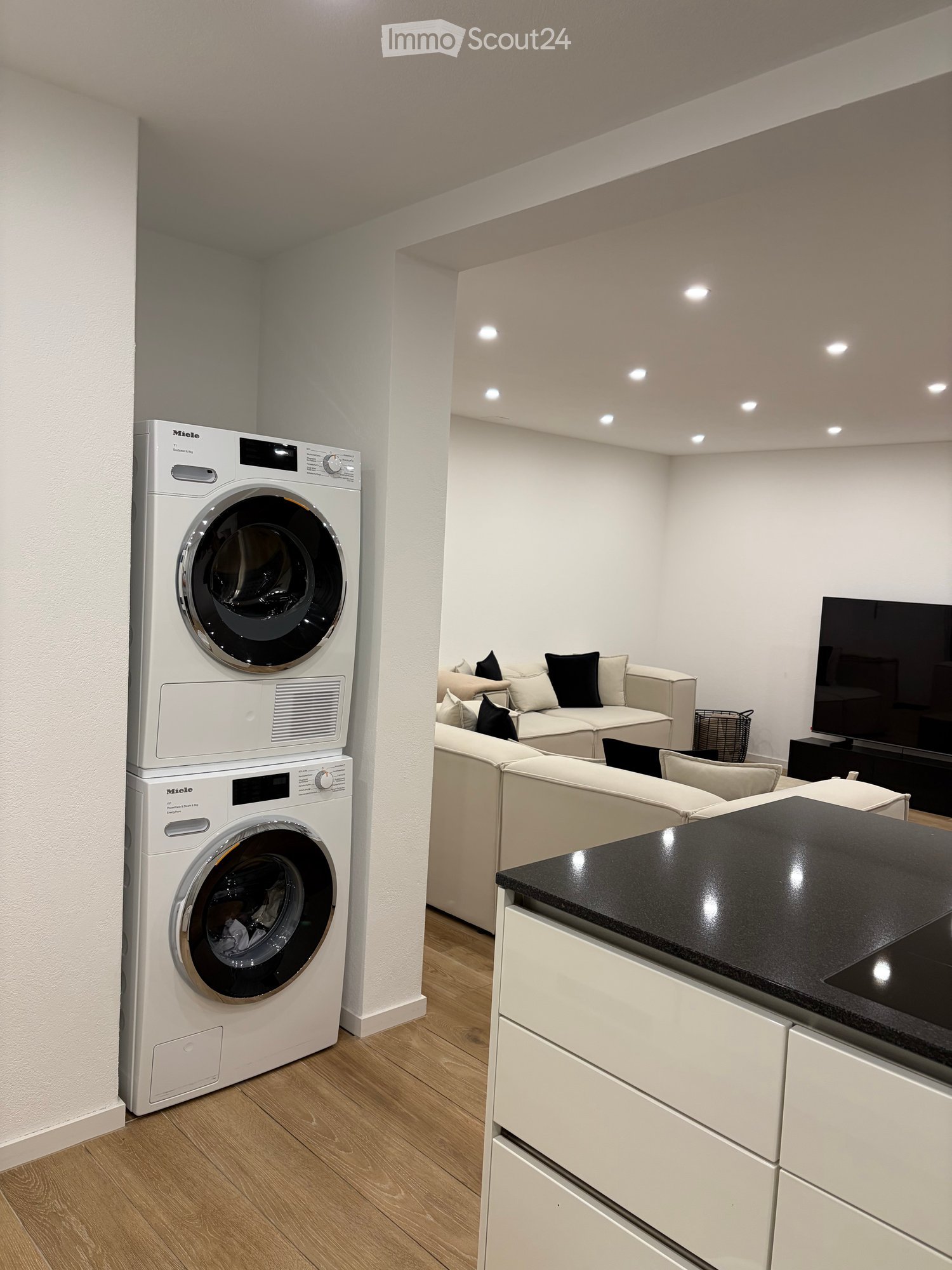 Modern living space with a white sofa, flat-screen TV, and a private washing machine unit. The room features light wooden flooring and white walls.