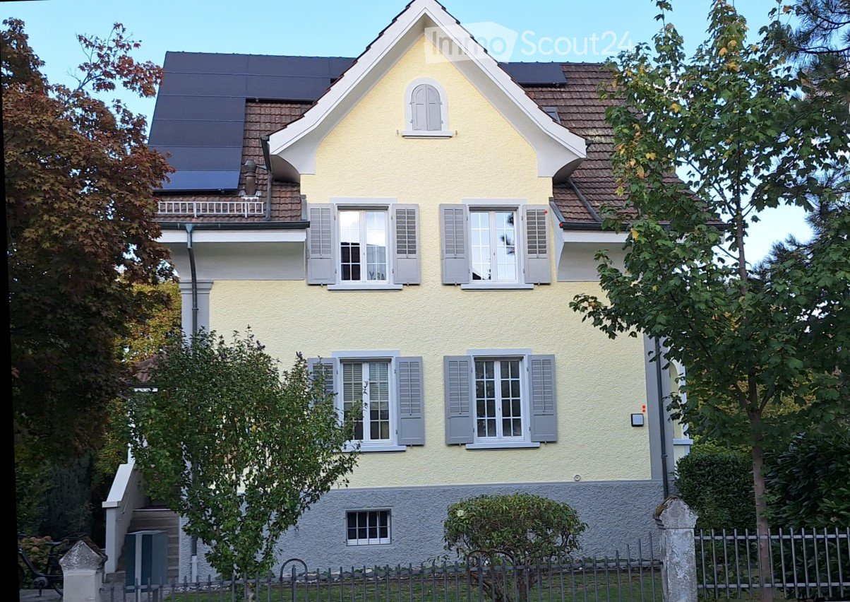 Two-story yellow house with white shutters, multiple windows, and a small balcony on the upper floor. The house features a gray stone base and is surrounded by trees and a metal fence.