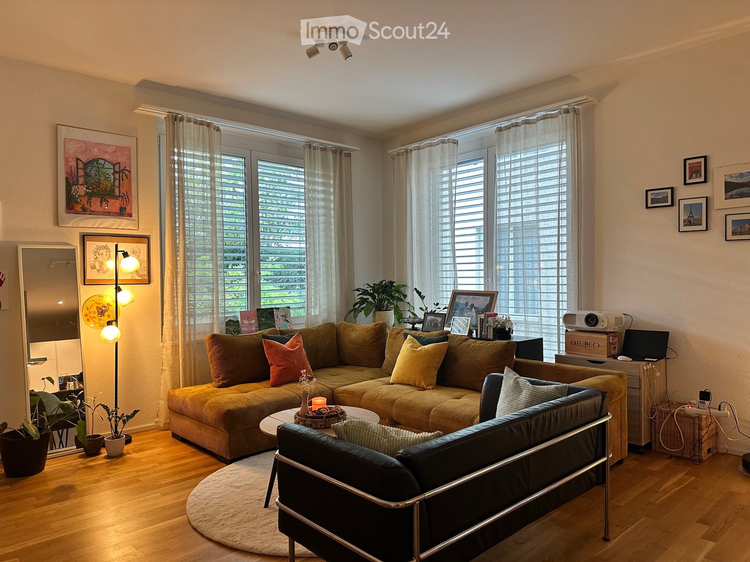 Bright living room with large windows, wooden flooring, a mustard-colored sectional sofa, a black leather armchair, a coffee table, a floor lamp, and a projector on a side table.