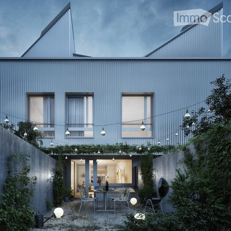 Modern two-story house with a sleek, grey exterior and large windows. The outdoor area features a small patio with a dining table, chairs, and string lights, surrounded by greenery and plants.