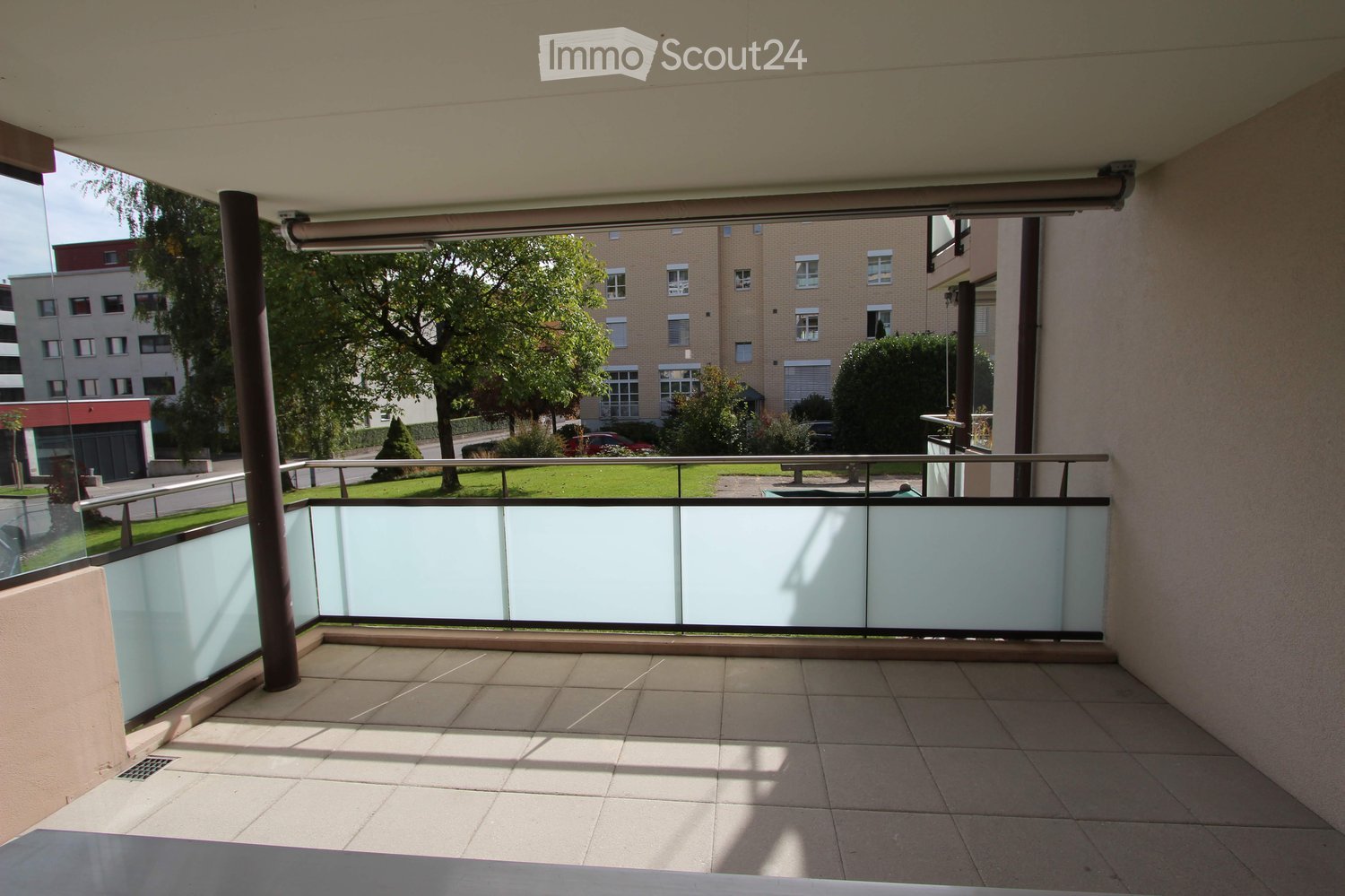Balcony with tiled floor, glass railing, and view of a green area with trees and buildings.