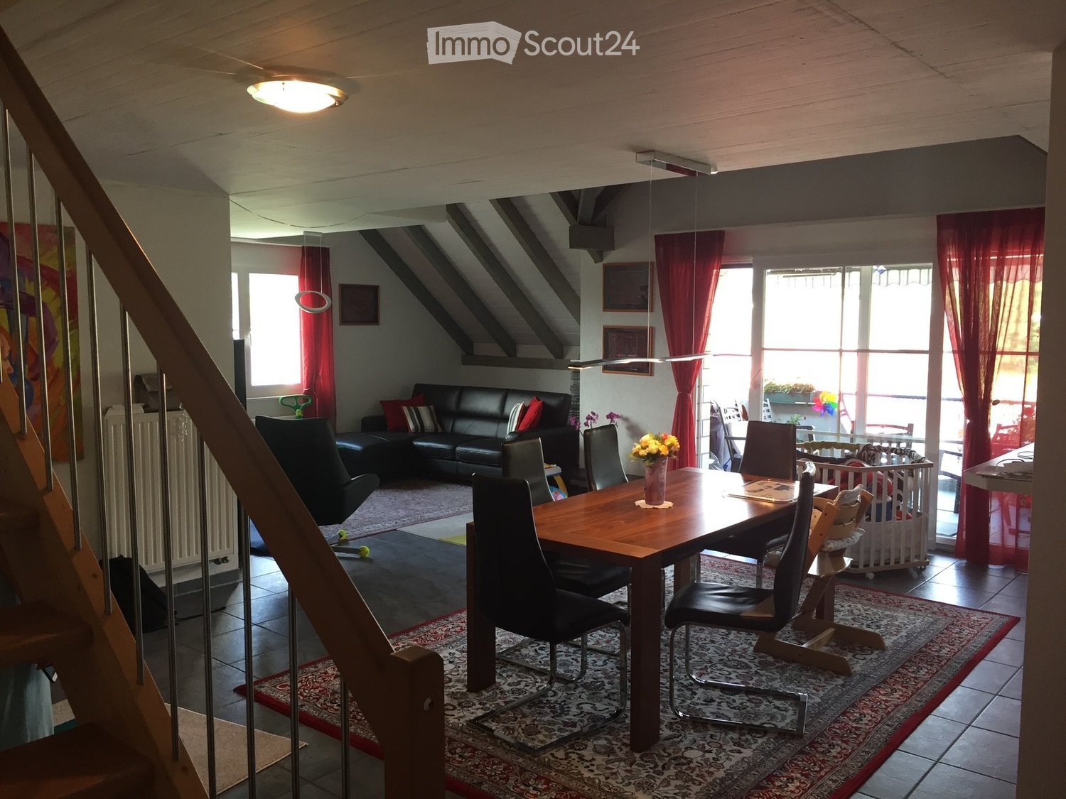 A well-lit living and dining area with a black leather sofa, a wooden dining table with chairs, a flower vase on the table, a baby crib, and large windows with red curtains.