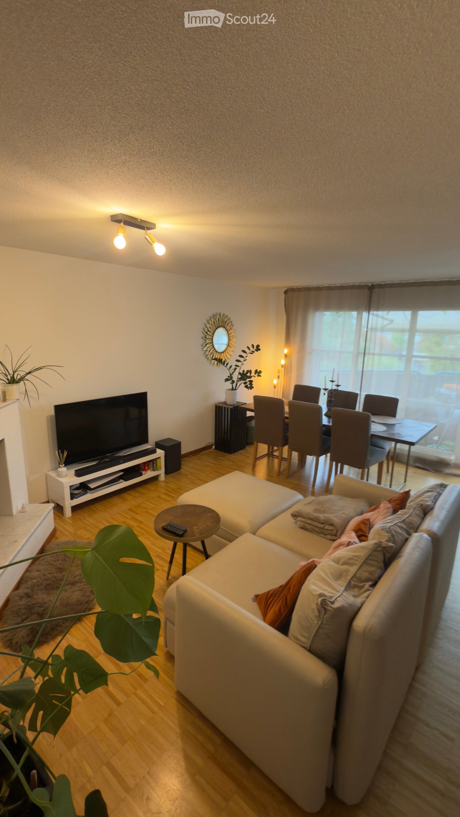 A modern living room with a beige sectional sofa, coffee table, TV, dining area with table and chairs, and decorative elements like a mirror and plants.