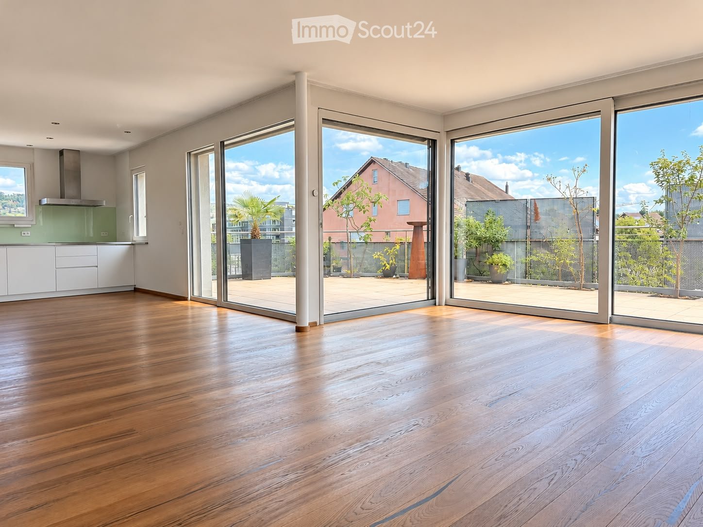 Bright, empty room with wooden flooring, large sliding glass doors leading to a balcony, and a modern kitchen area visible in the background.