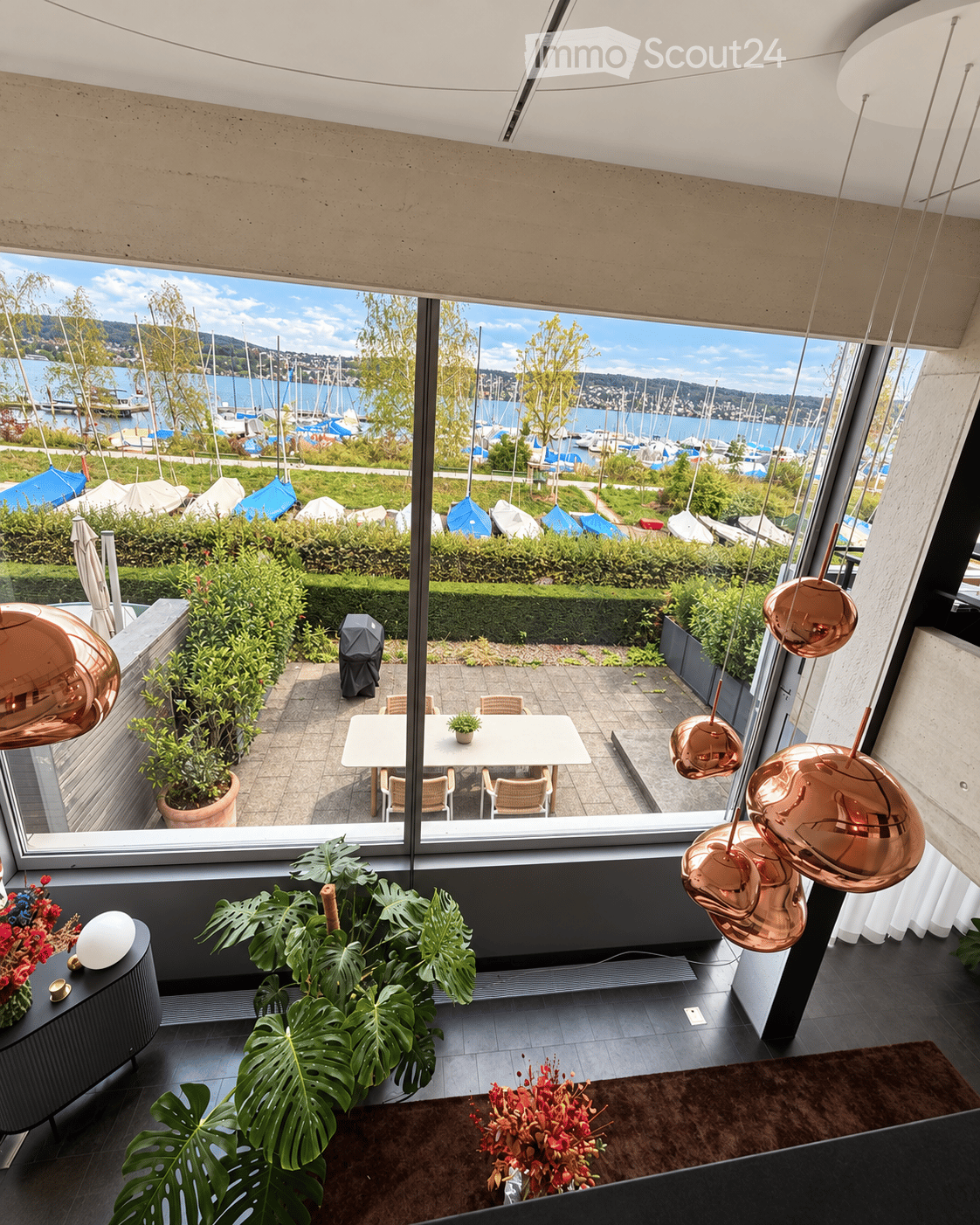 Modern interior with large windows offering a scenic view of a harbor and boats. Features a white dining table with chairs, potted plants, a flower arrangement, and decorative lamps.