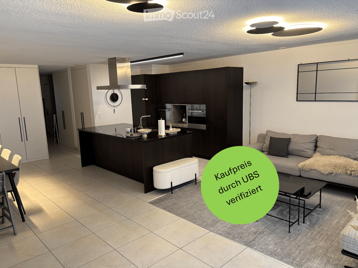 Modern open-plan living and dining area with a grey sofa, coffee table, bench, dining table, chairs, and a kitchen island with a faucet, oven, and cabinets. A large mirror and clock are mounted on the wall, and a floor lamp is present.