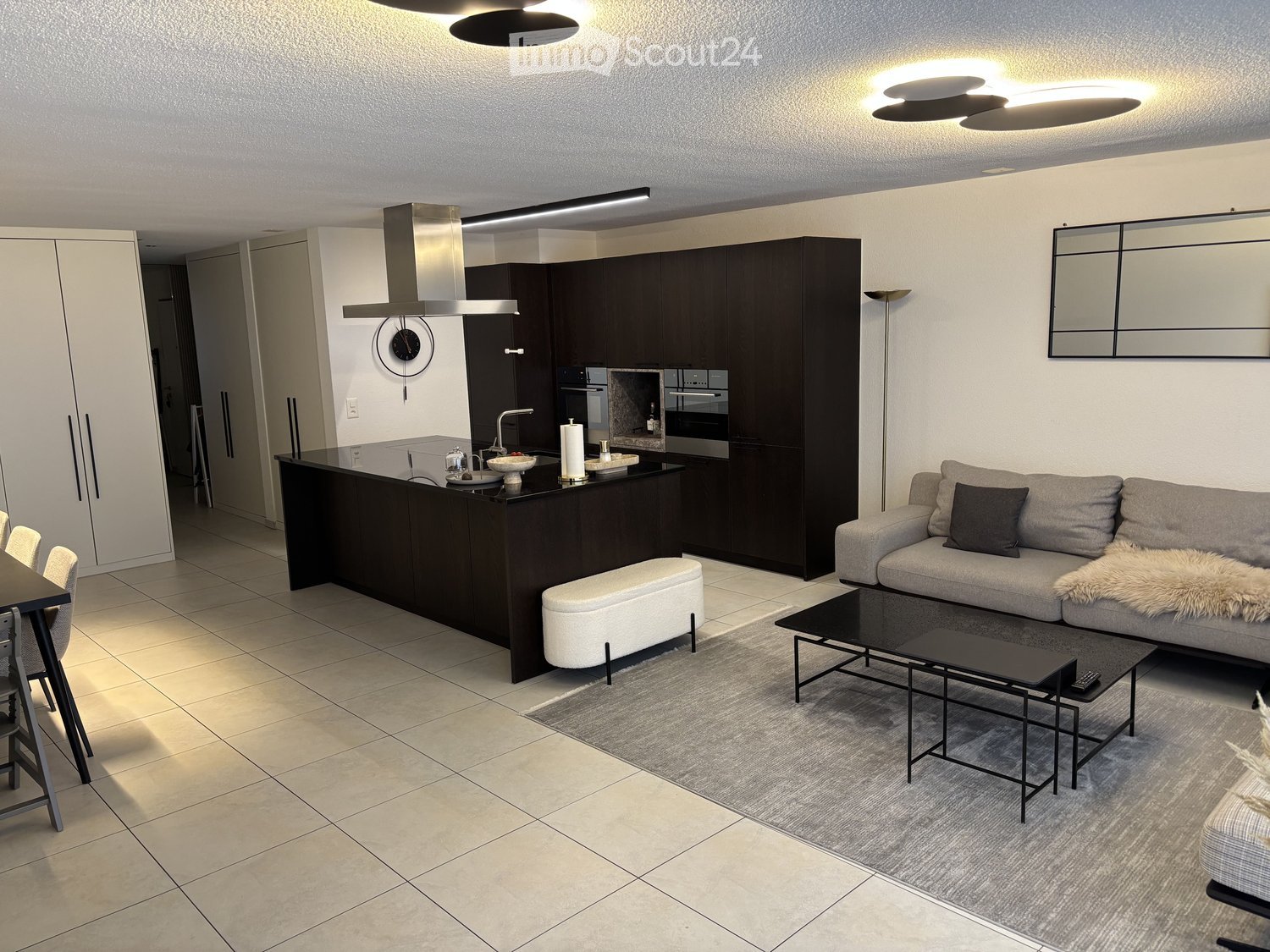Modern open-plan living and dining area with a grey sofa, coffee table, bench, dining table, and chairs. The kitchen features dark cabinets, a stainless steel range hood, and a central island with a sink and faucet. A large mirror and clock are mounted on
