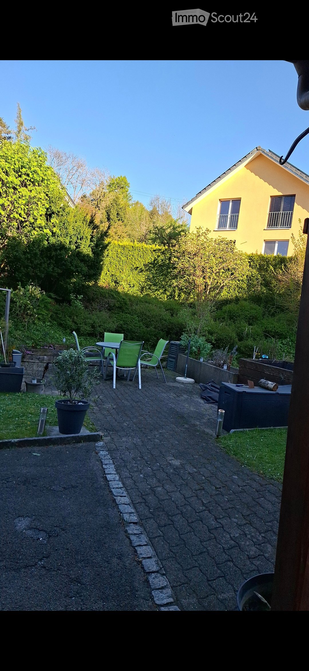 Garden area with a paved pathway, small seating area with a table and chairs, potted plants, and a yellow house in the background.