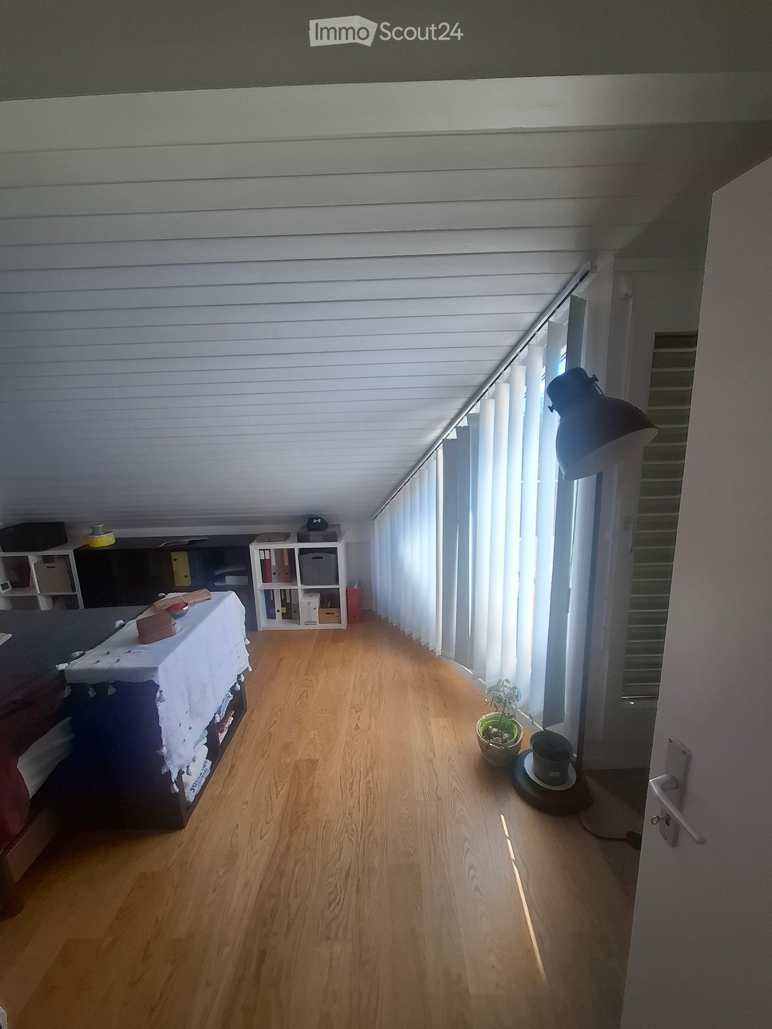 A furnished room with wooden flooring, a bed with a white tablecloth, a shelf with books and storage boxes, a lamp, a small plant, and a cup on the floor.