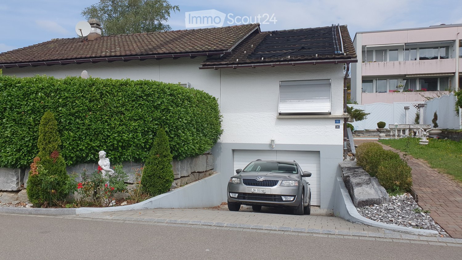 Two-story house with a tiled roof, white exterior, garage, and a parked car in front. The house is surrounded by greenery, including a hedge and small garden area.