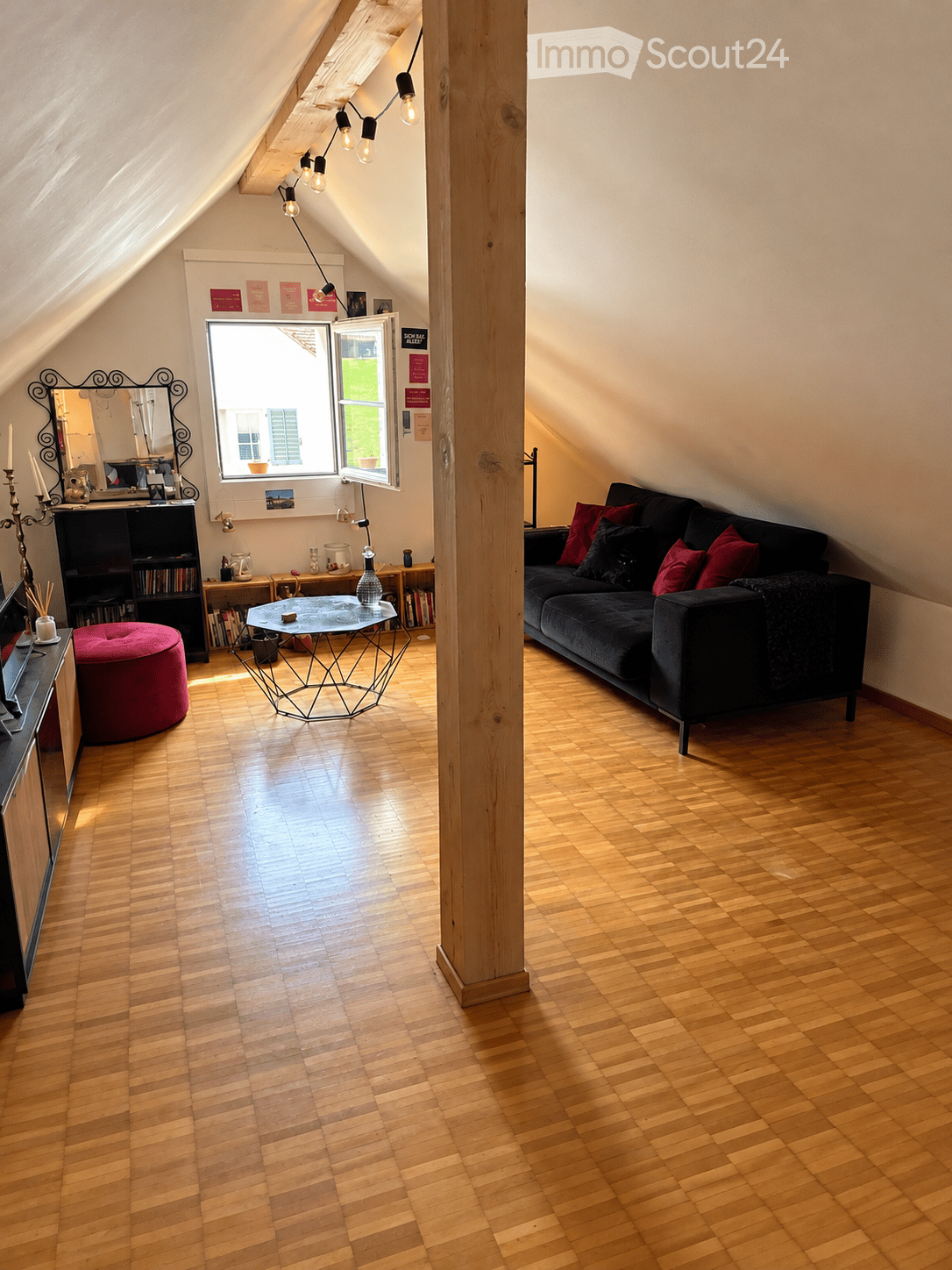 Attic living room with wooden flooring, slanted ceilings, and large windows. Features a dark-colored sofa with red pillows, a geometric coffee table, a red round stool, a bookshelf, and a mirror on the wall.