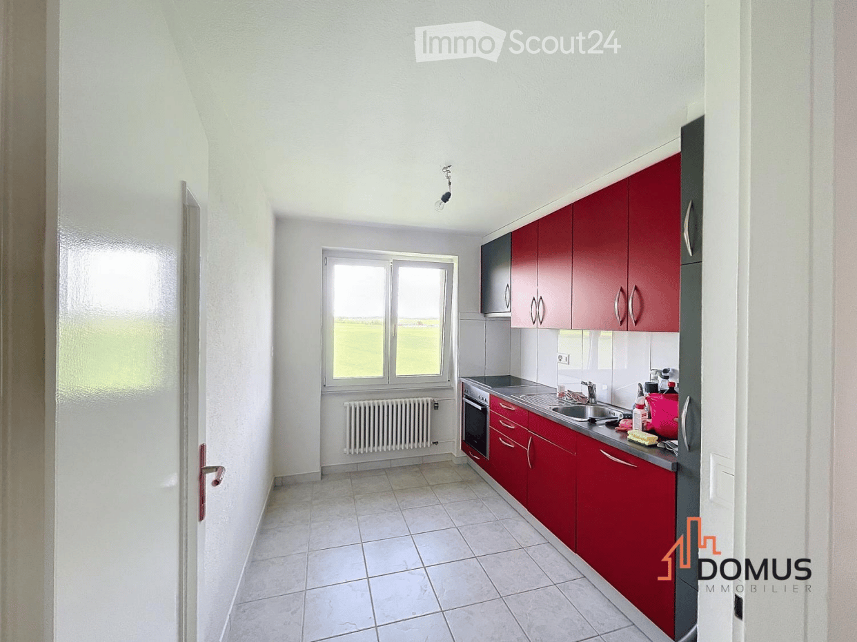 Modern kitchen with red cabinets, white tiled floor, and a window overlooking a green field. Includes a sink, oven, and radiator.