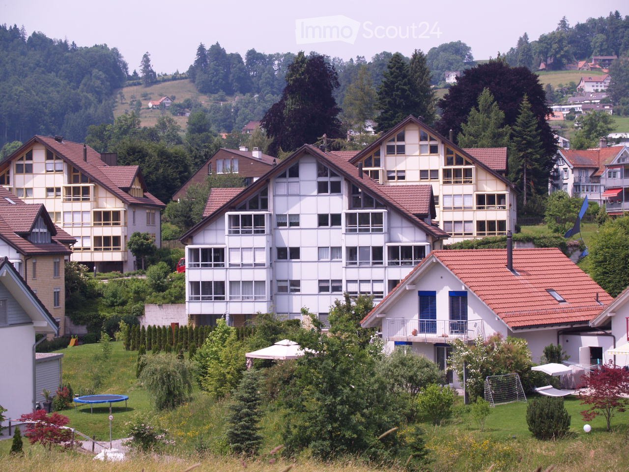 Residential area with multiple houses, surrounded by greenery and trees, with a mix of architectural styles and a scenic backdrop of hills.