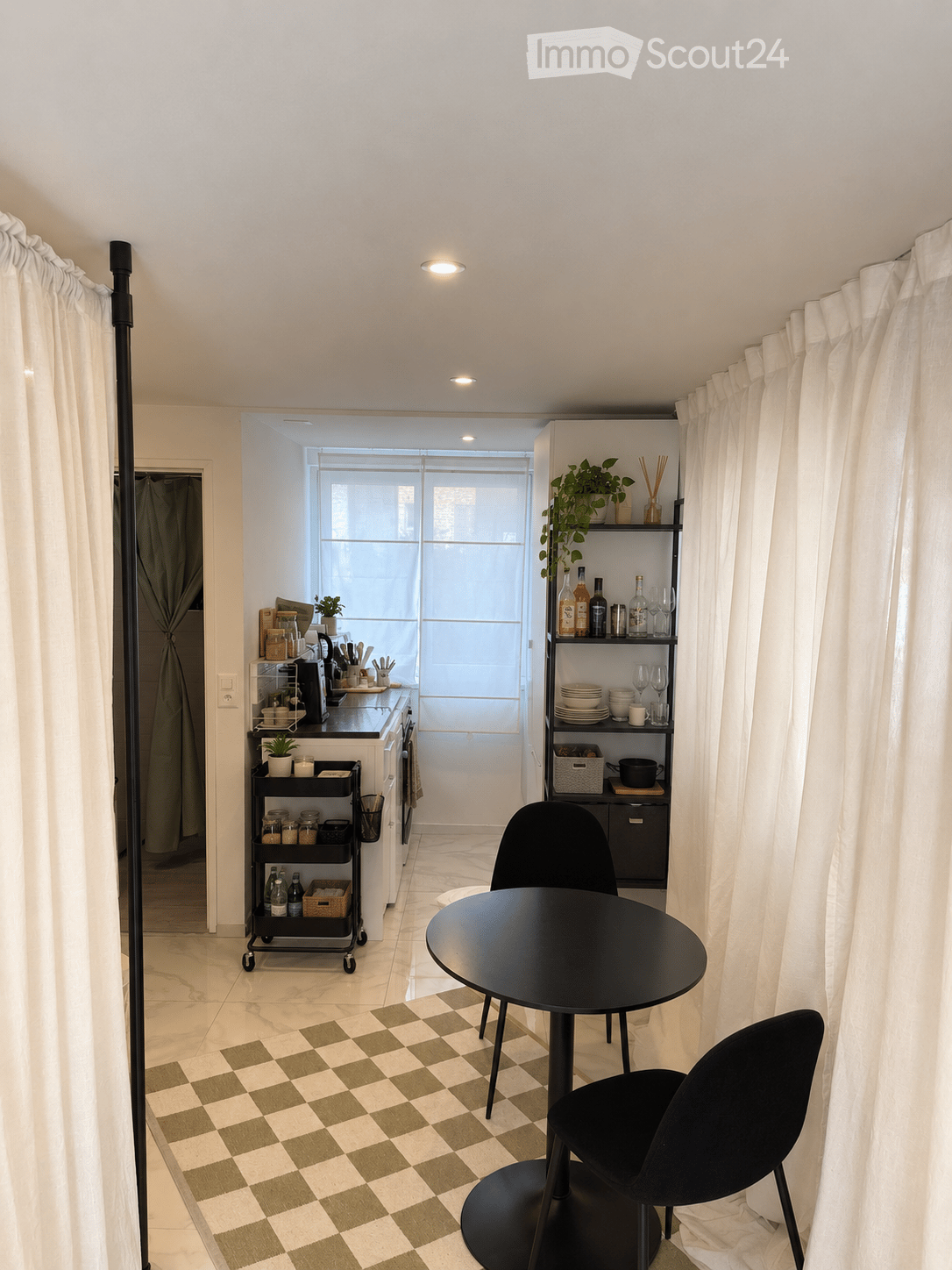 A modern, compact living space with a round black dining table and two chairs, a small kitchen area with a cart, shelves, and a window with blinds.