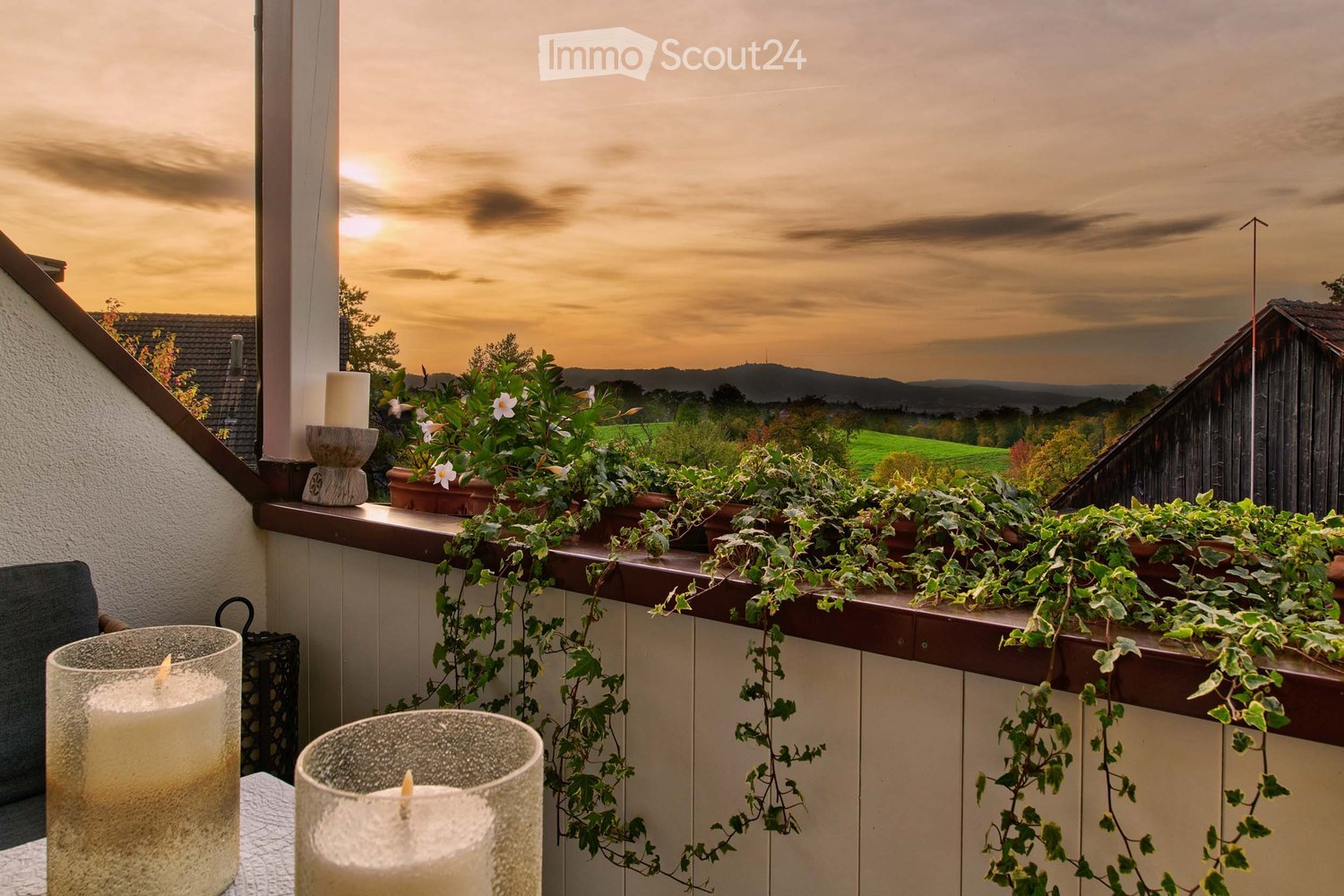 Balcony with potted plants, candles, and a scenic view of a countryside landscape at sunset.