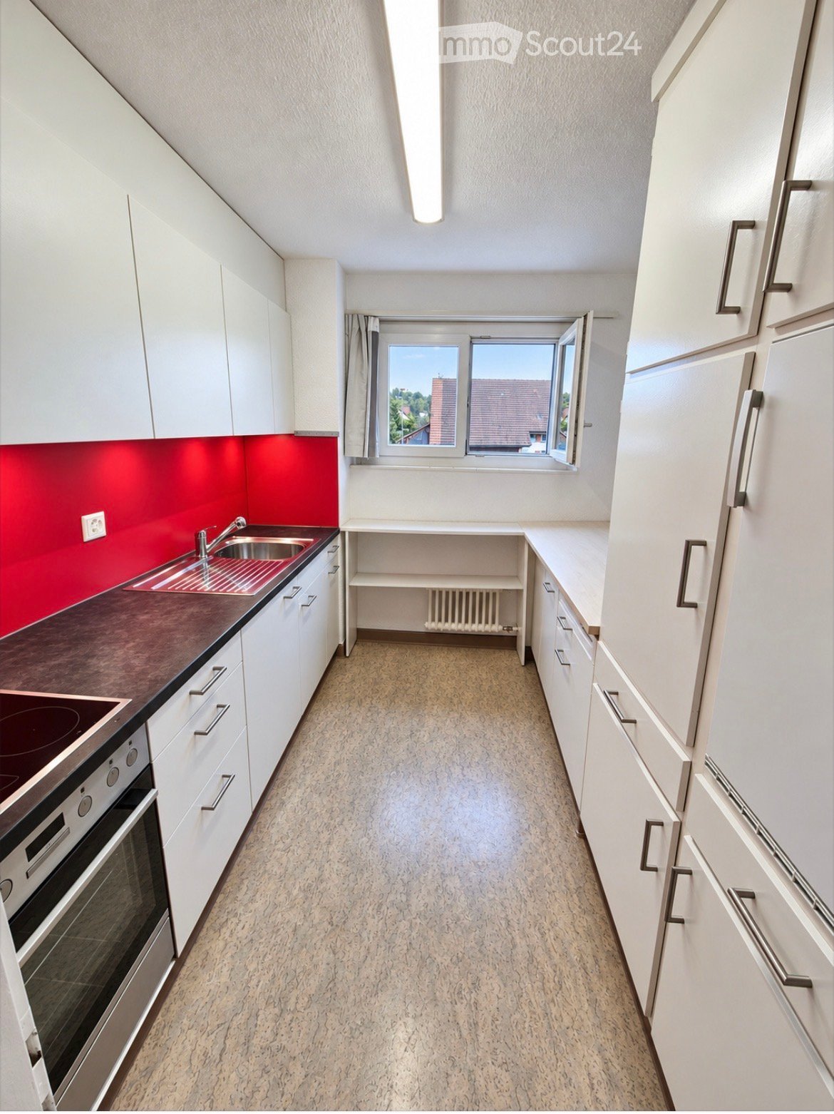 Modern kitchen with white cabinets, dark countertops, stainless steel sink, and built-in oven. Features a window with a view of the outside and a radiator below.