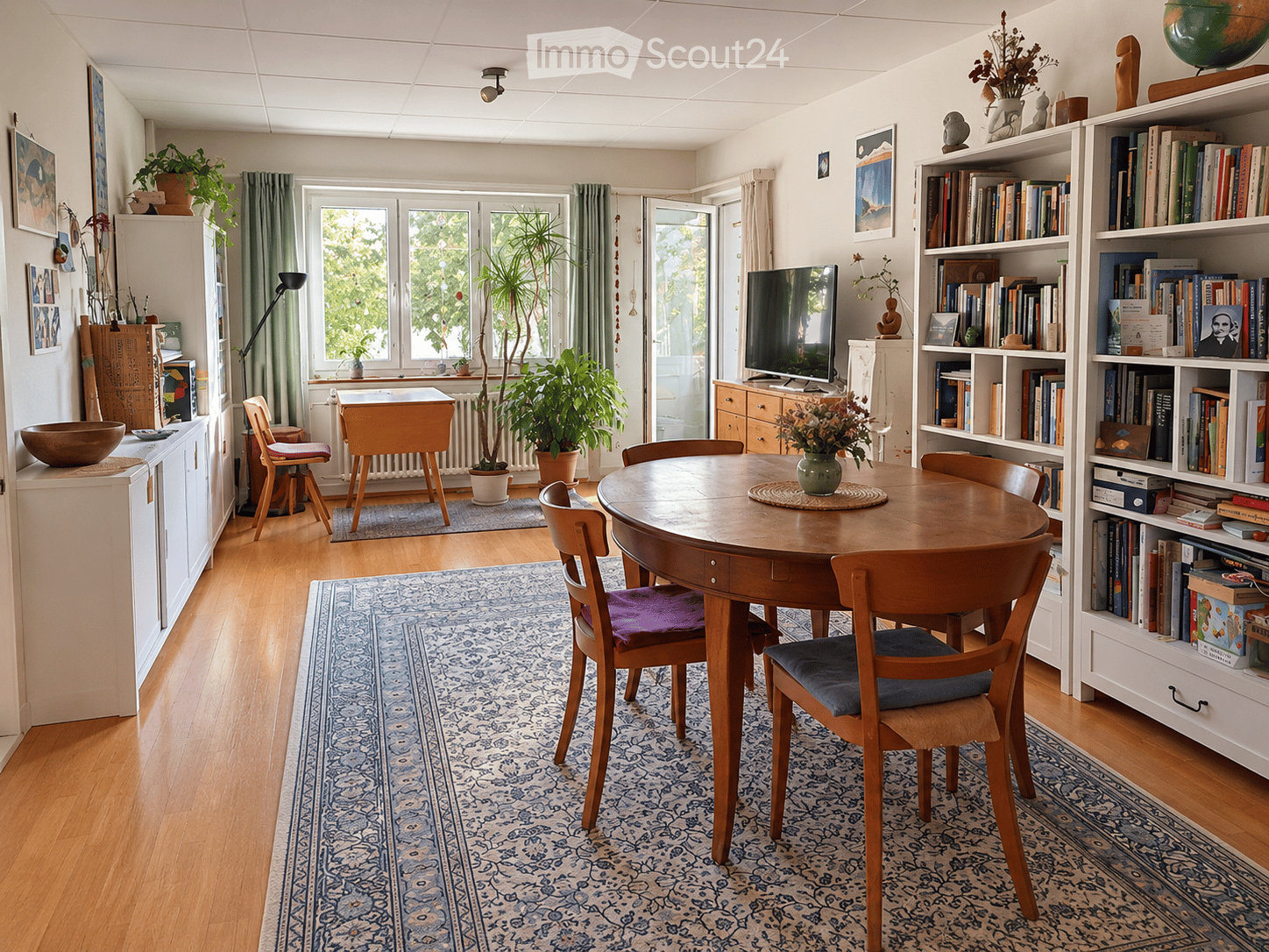 Brightly lit room with wooden flooring, a round wooden dining table surrounded by chairs, a bookshelf filled with books, a television, potted plants, and decorative items.