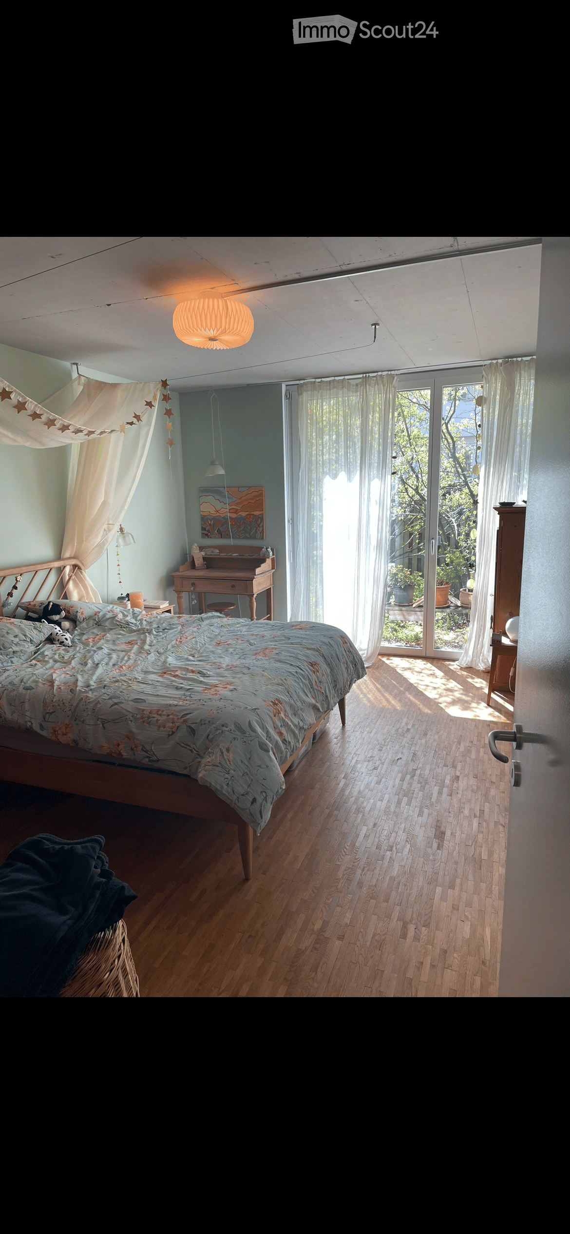 Bright bedroom with wooden flooring, large window with sheer curtains, bed with floral duvet, nightstand, desk, and artwork on the wall.