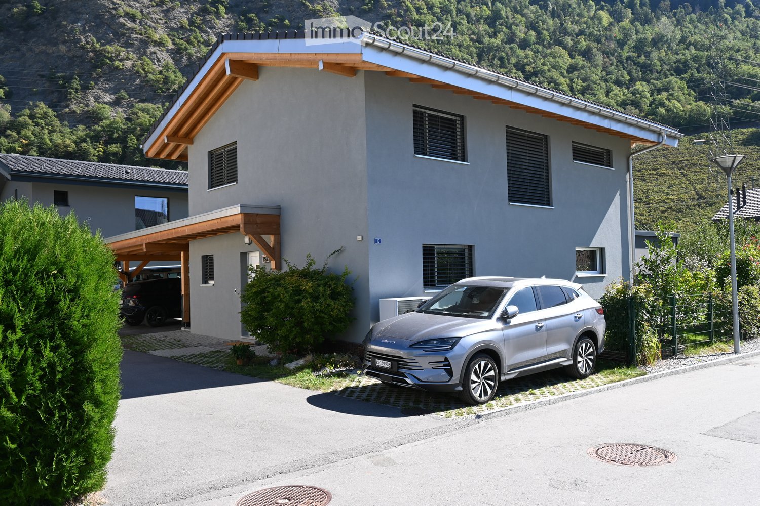 Modern two-story house with grey facade, wooden roof overhang, multiple windows, and a covered driveway with a parked SUV.