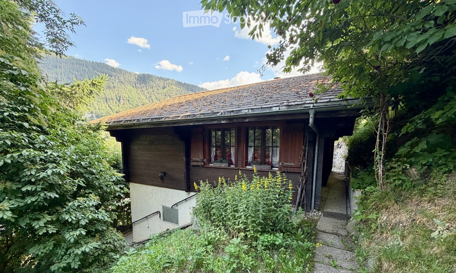 A small wooden house with a sloped roof, surrounded by lush greenery and trees, with a pathway leading to the entrance.