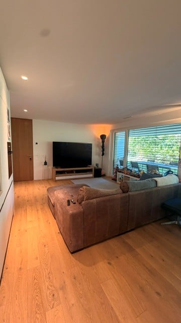 Modern living room with light wooden flooring, large windows offering a view, a grey sectional sofa, a coffee table, a television on a stand, and a floor lamp.