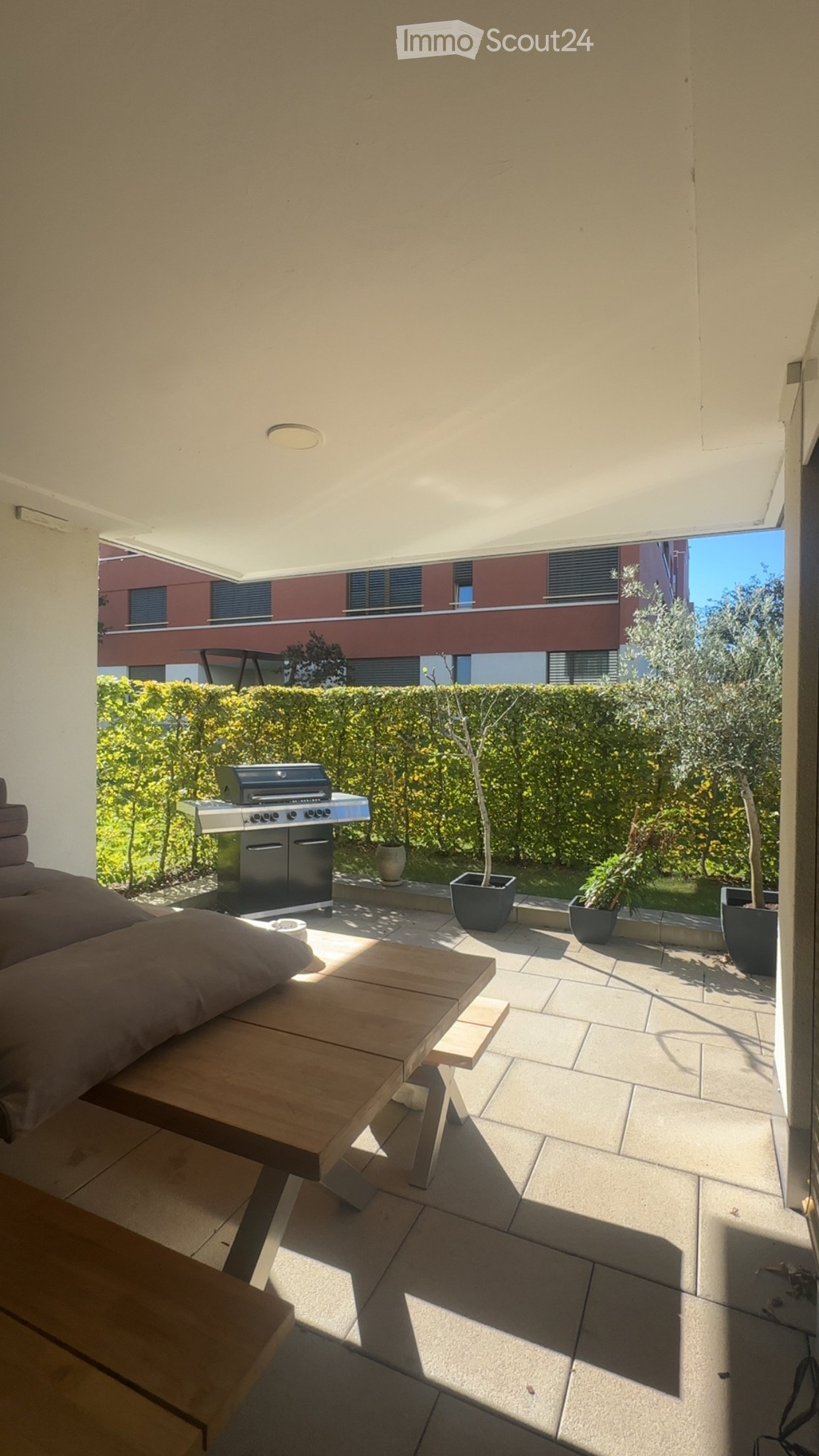 Balcony with a wooden table, sofa, grill, and several potted plants, overlooking a landscaped garden area.