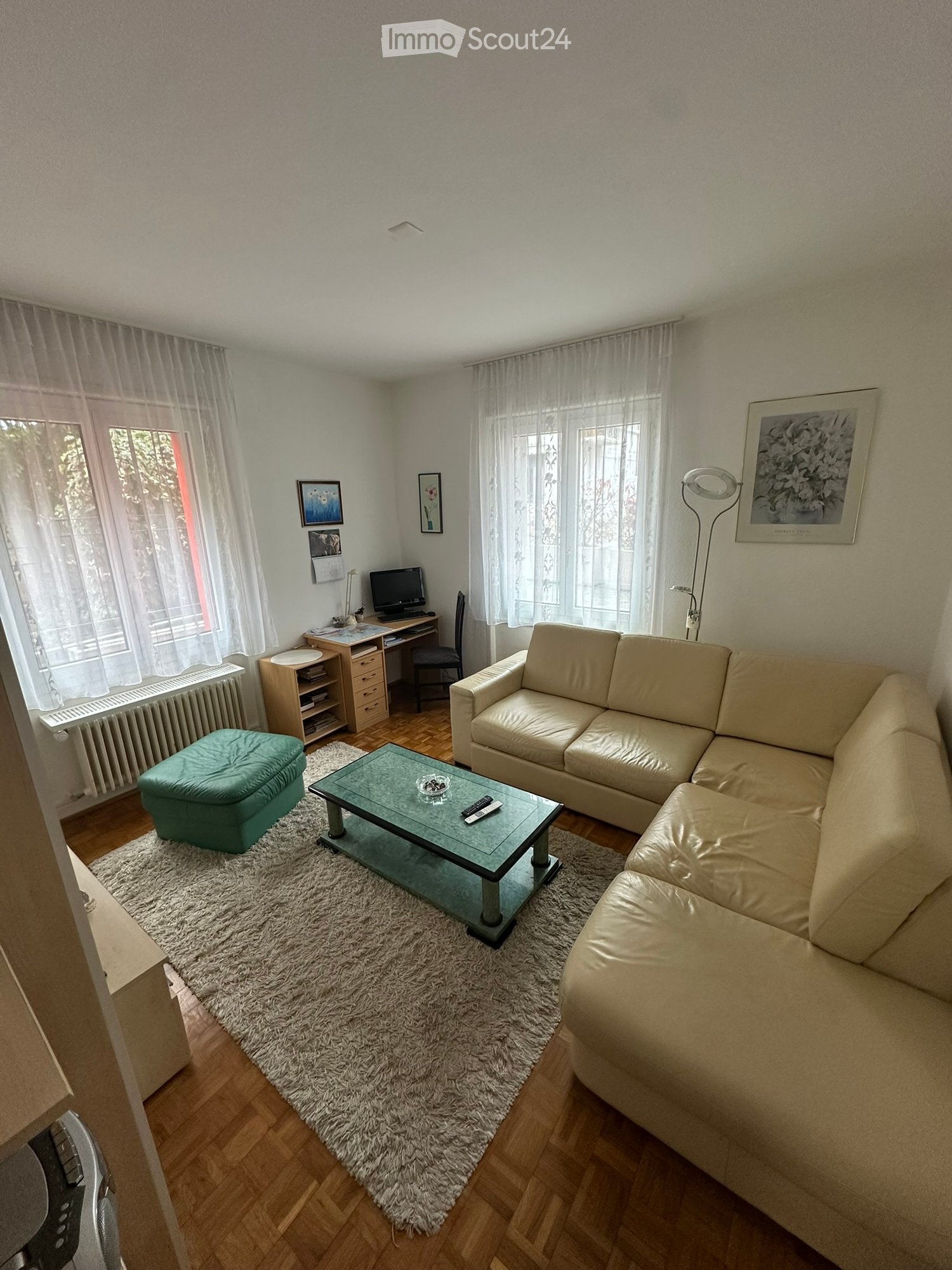 Living room with a cream-colored sofa, glass coffee table, teal ottoman, and a shaggy white rug on a wooden floor. A desk with a computer is positioned near a window with sheer curtains.