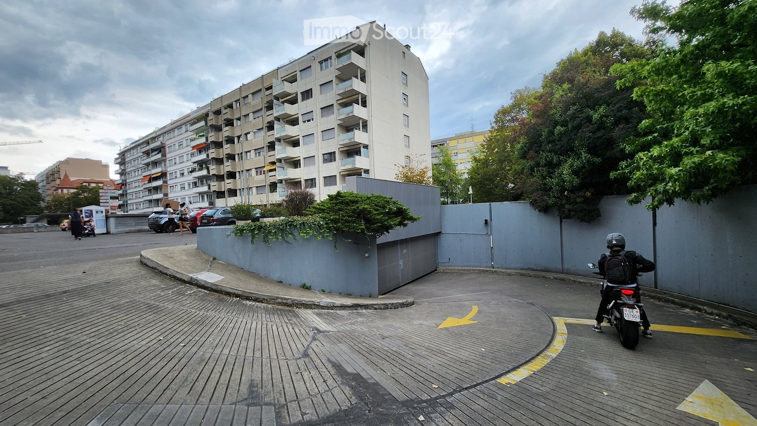 Multi-story apartment building with balconies, surrounded by trees and a paved area with a motorcycle passing by.
