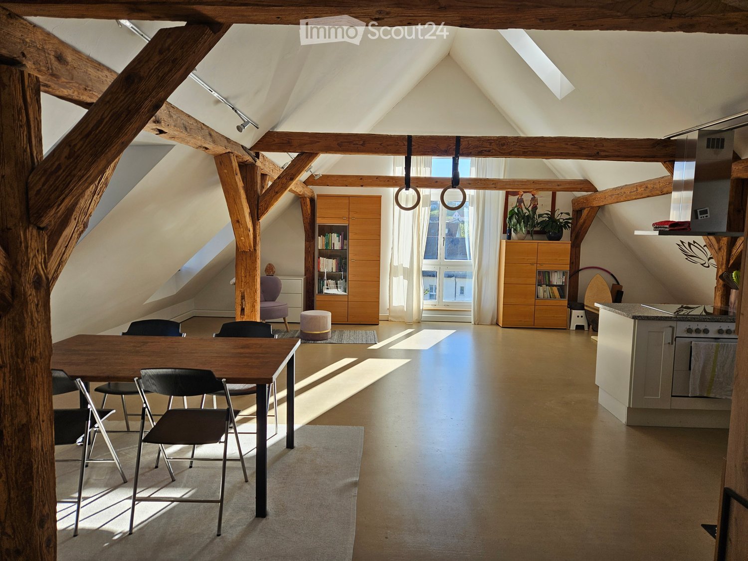 Open-plan attic space with wooden beams, a dining area with a table and chairs, a kitchen area, and built-in wooden cabinets. Natural light streams in from large windows.
