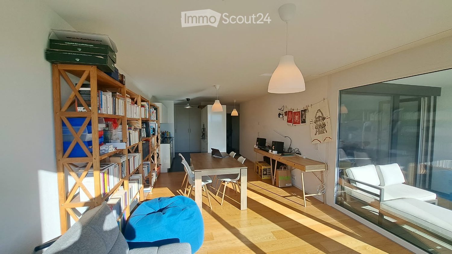 Bright, open-plan living and dining area with wooden flooring, large windows, a sofa, a blue ball chair, a dining table with chairs, a desk with a laptop, and a bookshelf filled with books.