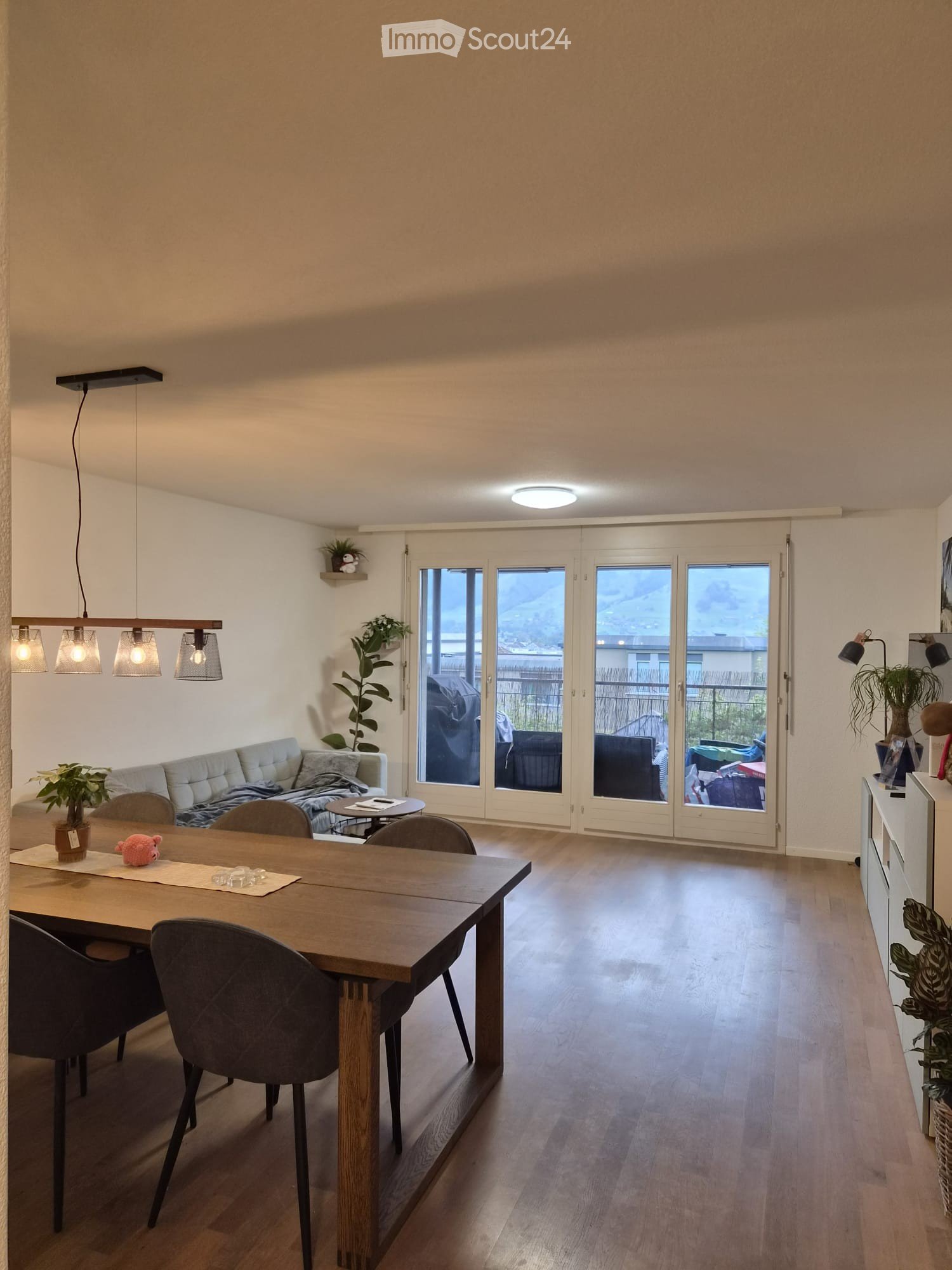 Bright living and dining area with wooden flooring, large sliding glass doors leading to a balcony, a sofa, dining table with chairs, pendant lights, and plants.