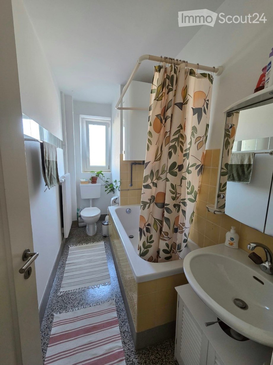 Bathroom with a bathtub, toilet, sink, mirror, and a shower curtain with a leaf and fruit design. The room features a small window, a towel on the wall, and a small plant on the floor.