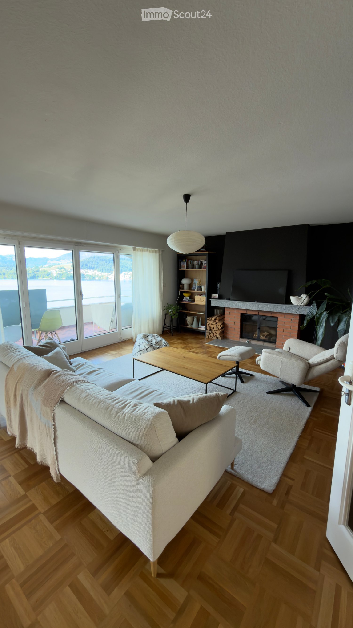 Modern living room with wooden flooring, large windows offering a scenic view, a beige sofa, coffee table, fireplace, bookshelf, and a television.