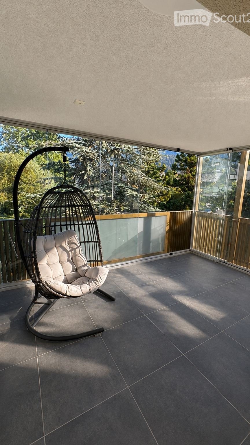 Balcony with tiled floor, glass railing, and a single hanging wicker chair with white cushions.