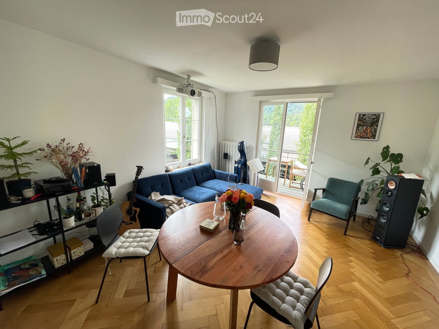 A modern living room with wooden flooring, a blue sofa, a round wooden table, chairs, a guitar, speakers, and plants. There is a large window and a sliding glass door leading to a balcony.