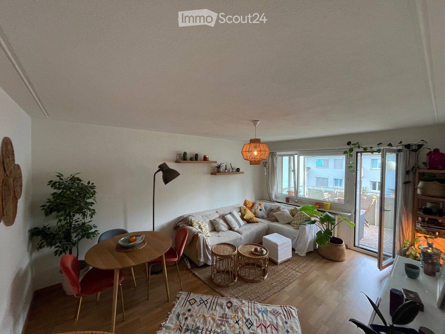 Bright living room with a white sofa, wooden dining table, colorful chairs, and a large window leading to a balcony. Features include a hanging lamp, floor lamp, and several plants.