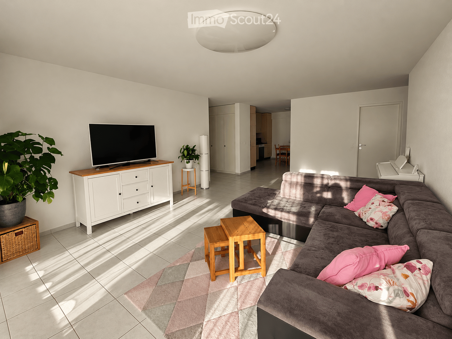 Modern living room with a grey sectional sofa, pink throw pillows, a coffee table, a TV on a white cabinet, and a large plant in a pot. The room features tiled flooring and natural light streaming in.